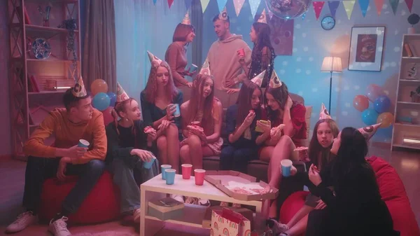 在一个家庭派对上 戴着节日礼帽的青少年们吃披萨 喝纸杯 这个女孩在电话录像中向她的朋友们展示 房间的内部装饰着一个迪斯科舞厅的宴会 — 图库照片
