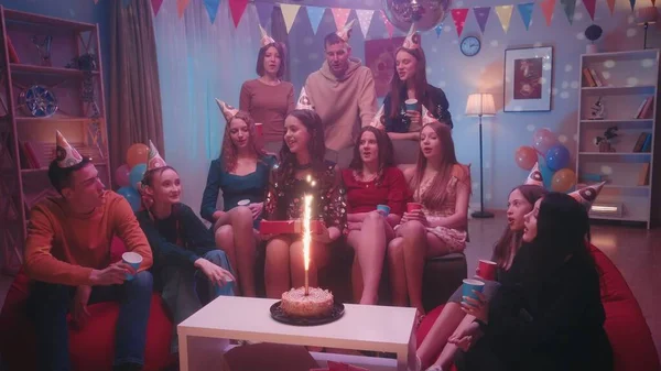 戴着节日礼帽的朋友祝贺生日的女孩 在为宴会布置的房间里 桌上放着一个点着烟火的蛋糕 在家里庆祝生日的概念 — 图库照片