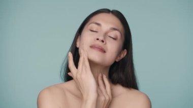 Çekici genç bir kadın parmaklarını yüzünde gezdiriyor, pürüzsüz ve sağlıklı bir cilde dokunarak. Mavi arka planda bir kadının portresi. Doğal güzellik, kozmetoloji, cilt bakımı. Yavaş çekim