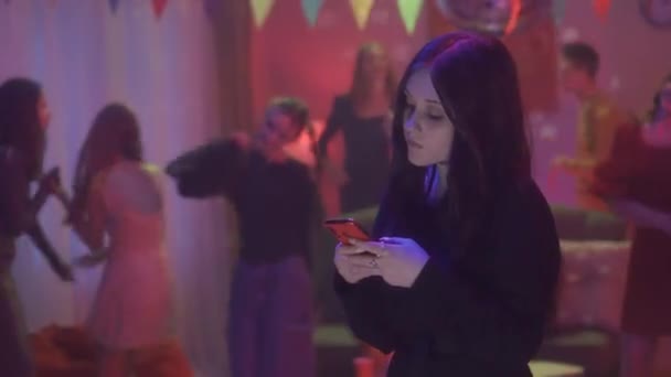 電話でビデオを見ているゴスの女の子は メッセージを入力し 彼女の友人がパーティーダンスで楽しんでいる間 ソーシャルメディアをチェックします 部屋はミラーディスコ付きの家パーティーのためにスマートに装飾されています — ストック動画