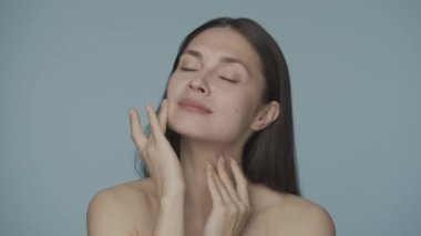Çekici genç bir kadın parmaklarını yüzünde gezdiriyor, pürüzsüz ve sağlıklı bir cilde dokunarak. Mavi arka planda bir kadının portresi. Doğal güzellik, kozmetoloji, cilt bakımı. HDR BT2020