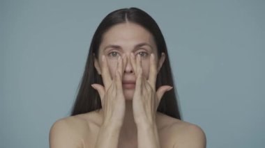 Elbiseleri olmayan genç esmer kadın yüzünün pürüzsüz ve sağlıklı cildine dokunuyor. Mavi arka planda izole edilmiş genç bir kadının portresi. Güzellik konsepti. Doğal güzellik, kozmetoloji, ten.