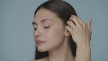 Genç bir kadın elini şakağından boynuna kadar uzatıyor, pürüzsüz, mükemmel tenini gösteriyor. Kadınların yüzleri mavi arka planda. Doğal güzellik kavramı, estetik cerrahi, kozmetoloji.