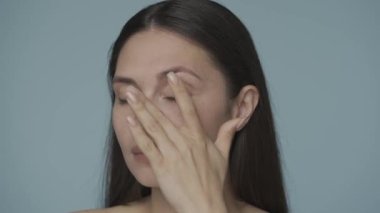 Bir seminer kadını göz çevresindeki deriye masaj yapar. Bir kadın, gözlerinin altındaki deriye nazikçe masaj yaparak kırışıklıklarla mücadele eder. Mavi arka planda bir kadın. Yakın plan. Doğal güzellik, yüz bakımı