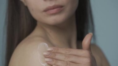 Yavaş masaj hareketleriyle, kadın kremi omzuna sürüyor. Bir seminer kadını cildine dikkat eder. Bulanık mavi arka planda. Cilt ve vücut bakımı kavramı. Doğal.