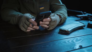 Bir Ukraynalı asker, üzerinde askeri silahı ve mühimmatı bulunan tahta bir masada oturur. Akıllı telefon tutan erkeklerin ellerini kapatıyoruz. Bir asker aile fotoğrafına bakar, bir mesaj yazar ve konuşur.