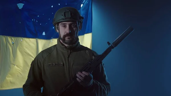 カラシニコフのアサルトライフルでウクライナ軍の男を閉じます 兵士は暗闇の中で弾丸の穴でいっぱいのウクライナの旗の背景に立って サーチライトのビーム — ストック写真
