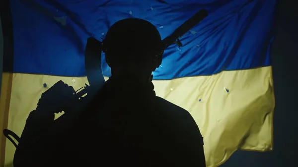 弾丸の穴に覆われたウクライナの旗を背景に ウクライナの兵士のシルエットのクローズアップ 兵士はスポットライトの暗闇の中に立って 肩にライフルを持っている — ストック写真