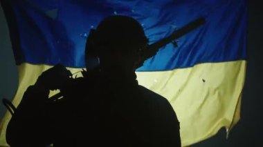 Ukrayna bayrağının arka planında kurşun delikleriyle kaplı bir Ukrayna askerinin siluetini kapat. Bir asker karanlıkta, spot ışıklarının altında tüfeğini omzuna dayar. Yavaş