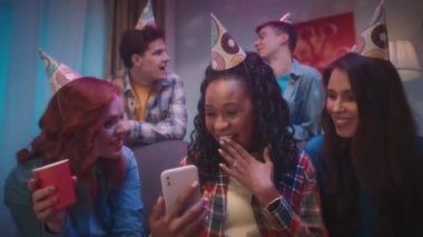 Afrikalı bir Amerikalı kız telefonunu arkadaşlarına gösterir. Duygusal olarak fotoğraflara, videolara ya da video konuşmalarına bakarlar. Parti şapkalı bir grup arkadaş parti için dekore edilmiş bir odada.