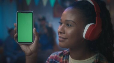Kafasında kırmızı kulaklık olan Afro-Amerikalı bir kız yeşil ekranlı bir telefon gösteriyor. Genç bir kız, bir odanın arka planında bulanık bir telefon tutuyor. Teknoloji, cihaz.