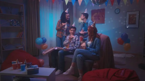 一群戴着派对帽的年轻人坐在房间的沙发上 一边吃披萨 一边喝酒 房间的内部装饰着迪斯科舞会 气球和纸旗 — 图库照片