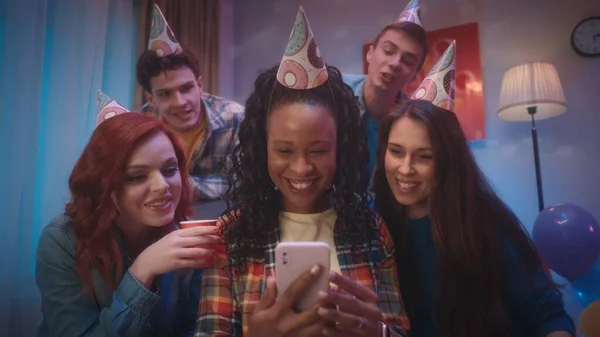 お祝いの帽子の若者たちは アフリカ系アメリカ人の女の子が持っている電話を見ています 友人はビデオ通話 共有写真やビデオを介して通信し 思い出に残る 古い友人 パーティー 誕生日に会う — ストック写真