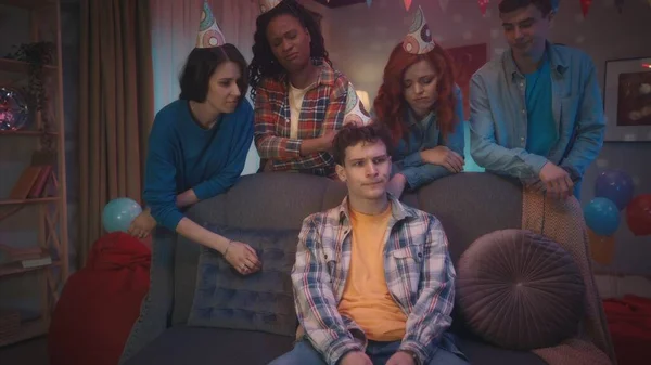 一个卷发的家伙 戴着派对帽 被他的朋友们簇拥着 坐在沙发上因为比赛的失败而心烦意乱 朋友们支持他 鼓励他 家庭伴侣的娱乐 — 图库照片