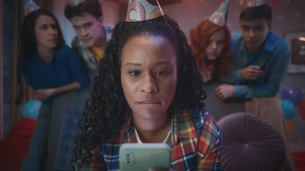 アフリカ系アメリカ人女性は緊張から彼女の唇をかむ 彼女の電話でオンラインゲームをプレイしています 女の子の友人は彼女の後ろに立って 彼女と心配しています お祝いの帽子をかぶった若者たちが — ストック写真