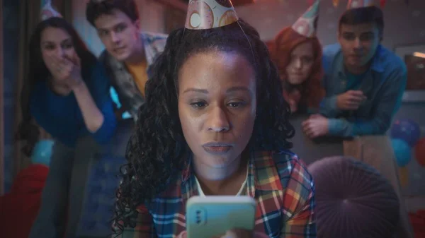 アフリカ系アメリカ人の女性がソファに座り 彼女の携帯電話 ビデオを見て ソーシャルネットワークを閲覧します 女の子の友達は彼女の後ろに立っている お祝いの帽子をかぶった若者のグループ — ストック写真