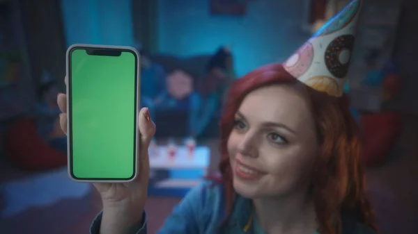 一个戴着节日礼帽的红头发女孩笑着展示了一个带着绿色屏幕的智能手机 在房间模糊的背景上 一个拿着智能手机的女孩的近身镜头 小玩意 广告概念 — 图库照片