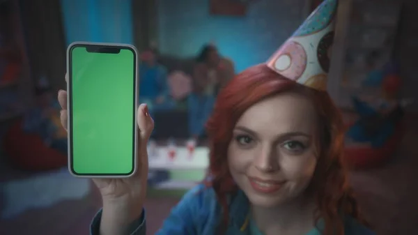 一个戴着节日礼帽的红头发女孩笑着展示了一个带着绿色屏幕的智能手机 在房间模糊的背景上 一个拿着智能手机的女孩的近身镜头 小玩意 广告概念 — 图库照片