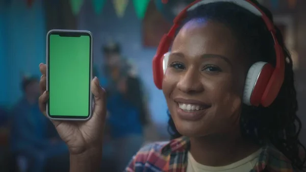 頭に赤いヘッドフォンをしたアフリカ系アメリカ人の女の子が緑色の画面を持つ携帯電話を示しています 部屋の中でパーティーのぼやけた背景に携帯電話を持っている若い女の子 — ストック写真