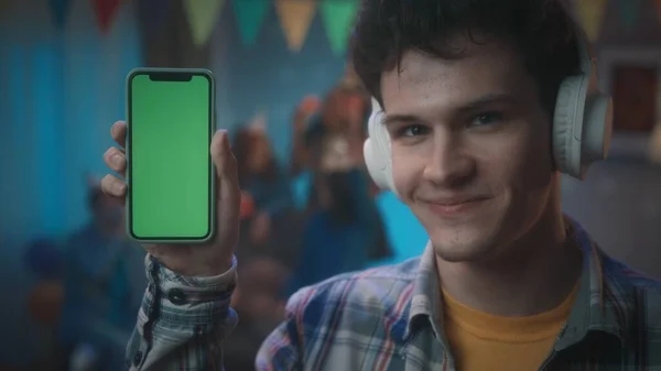 戴白色耳机的家伙展示了一个带有绿色屏幕的智能手机 在派对的模糊背景下 在一个装饰着迪斯科舞会和国旗的房间里 一个拿着智能手机的家伙的近照 — 图库照片