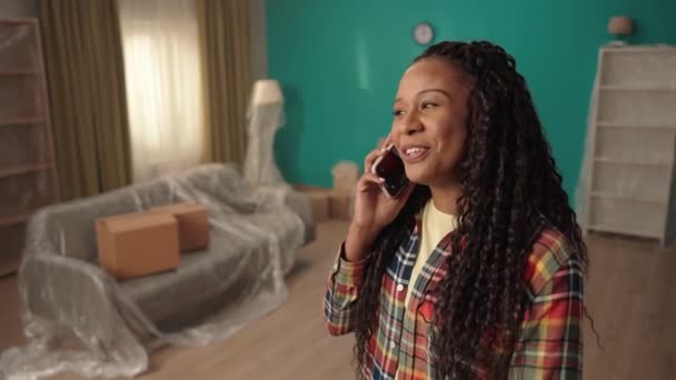 アフリカ系アメリカ人の女の子が喜んで電話で話し 新しいアパートのリビングルームに立っています 携帯電話を持つ少女の肖像画 不動産の購入 引越し 住宅温暖化 — ストック動画