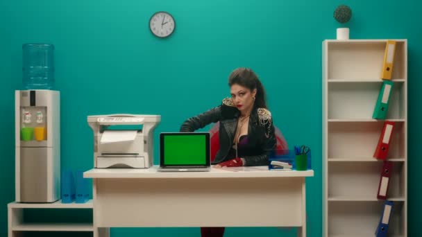 用食指指尖着绿色笔记本电脑屏幕微笑的女性怪胎走近了 一个妆容光彩夺目的女人的画像 穿着黑色皮夹克 带着锁链坐在办公室的桌子旁 — 图库视频影像