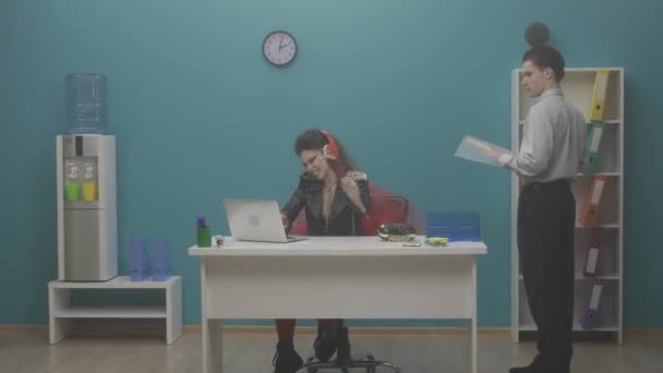 女性のフリークは ヘッドフォンでラップトップでオフィスに座って 音楽に耳を傾け 歌います 従業員は近くに立っていて 同僚を唖然とさせ 去っていきます 音楽アプリケーション — ストック動画