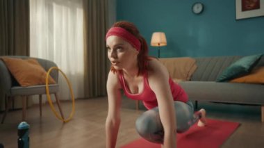 Pembe spor giysili kızıl saçlı genç kadın ısınıyor oturma odasında bir spor minderinin üzerinde geriniyor. Ev fitness konsepti