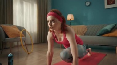 Pembe spor giysili kızıl saçlı genç kadın ısınıyor oturma odasında bir spor minderinin üzerinde geriniyor. Ev fitness konsepti