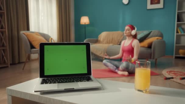 客厅的桌子上有带绿色屏风的笔记本电脑 在背景音乐中 一个戴耳机的女人坐在荷花的位置上 听着音乐进行冥想 您的文本模板位置 — 图库视频影像
