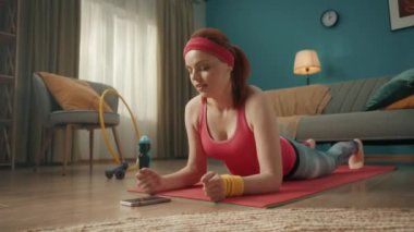 Spor giysili bir kadın telefonun önünde yerde yatıyor. Bir kadın telefonunda zamanlayıcı başlatır ve dirseklerinin üzerine tahta bir konuma gelir. Ev fitness konsepti, online spor sınıfı