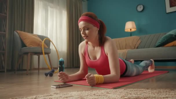 一名身穿运动服的妇女躺在电话前的地板上 一个女人在她的手机上开始计时器 并在她的手肘上进入一个木板的位置 家庭健身概念 在线体育课 — 图库视频影像