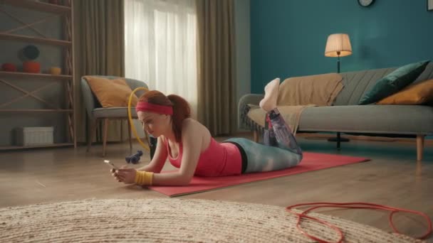 一位穿着粉色运动服的红头发年轻女子躺在客厅的地板上 一个女人手里拿着一部电话 填着一本电子培训日志 笑着对这一切感到满意 — 图库视频影像