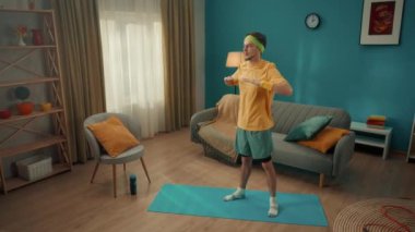 Evde spor haberleri. Sarı spor kıyafetli ve kafasında bandaj olan bir adam oturma odasında ısınıyor. Adam vücuduyla dönüşler yapıyor ve ellerini arkaya alıyor, kürek kemiklerini birleştiriyor.