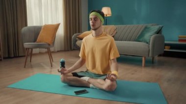 Spor giyimli genç bir adam oturma odasında akıllı telefonun yanında lotus pozisyonunda oturuyor. Meditasyon, rahatlama. Spor konsepti, ev antrenmanı.