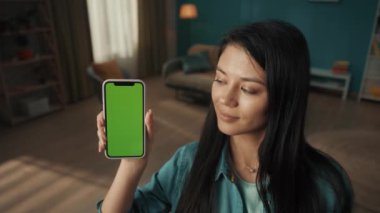 Genç gülümseyen kadın yeşil ekranlı akıllı telefon gösteriyor. Oturma odasında akıllı telefonlu Asyalı bir kadının portresi. Teknoloji, cihaz, reklam konsepti