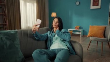 Çekici Asyalı kadın akıllı telefondan selfie çekiyor, poz veriyor, gülümsüyor. Oturma odasındaki kanepede oturan genç bir kadın elinde telefon var.