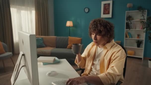 一个卷曲的小伙子坐在电脑前 喝着早晨的咖啡和茶 男人在键盘上打字 写电子邮件 上网工作 自由职业者 一个卷曲的年轻人的肖像特写 — 图库视频影像