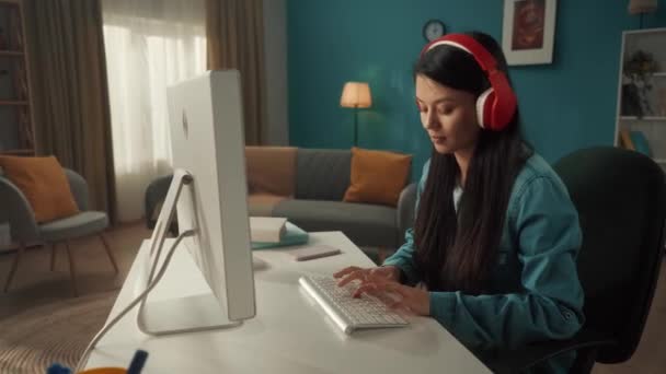 赤のワイヤレスヘッドフォンの女性はコンピュータで動作します 若い女性が音楽に耳を傾け 音楽のビートに移動し キーボードに入力します リビングルームのアジアの女性のプロフィール肖像画 — ストック動画