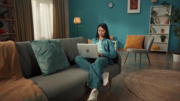 魅力的なアジアの女性は 彼女の膝の上にラップトップを持つリビングルームのソファに座っています 女性が働いている テキスト メッセージ チャットを入力しています リモートワークフリーランサー — ストック動画