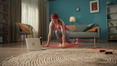 Spor kıyafetli kızıl saçlı genç bir kadın ısınıyor oturma odasında spor minderinin üzerinde geriniyor. Bir kadın dizüstü bilgisayarda video dersi izliyor. Ev fitness konsepti, online spor sınıfı. Yavaş