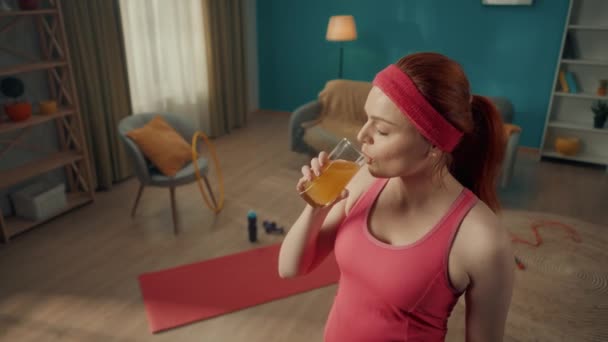 一个红头发的女人在运动后喝新鲜榨果汁的肖像 一个穿着运动服的女人 靠着客厅的后面 有训练的特征 杂乱的东西 — 图库视频影像