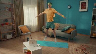 Oturma odasındaki paspasın üstünde yıldız atlayışı yapan genç adam. Bir adam dizüstü bilgisayardan video dersi izler ve egzersizleri tekrarlar. Ev fitness konsepti, online spor sınıfı. Yavaş çekim