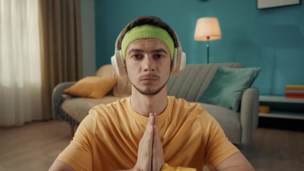 一个戴耳机的年轻人 听智能手机里的音乐 坐在莲花的位置 做瑜伽 一个穿着运动服的男子坐在客厅地板上的画像 — 图库视频影像