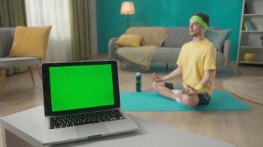 Yeşil ekranlı laptop oturma odasındaki masanın üzerinde. Adam nilüfer pozisyonunda oturuyor. Spor konsepti. Yoga dersi. Metnin için şablon yeri, promosyon içeriği. Reklam alanı