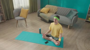 Spor giyim sektöründe genç bir adam oturma odasında, akıllı telefonun yanında Lotus pozisyonunda oturuyor. Meditasyon, rahatlama. Spor konsepti, ev antrenmanı. HDR BT2020 HLG Malzemesi
