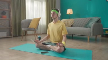 Spor giyimli genç bir adam oturma odasında akıllı telefonun yanında lotus pozisyonunda oturuyor. Bir adam meditasyon yapar, dinlenir, beyaz kablosuz kulaklıkla müzik dinler. Spor konsepti, ev