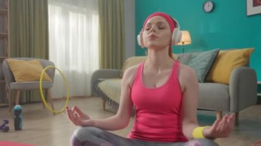 Kızıl saçlı, kulaklıklı genç bir kadın lotus pozisyonunda oturur, meditasyon yapar, yoga yapar. Spor kıyafetli bir kadının portresi oturma odasında yerde oturuyordu. Rahatlama, meditasyon.