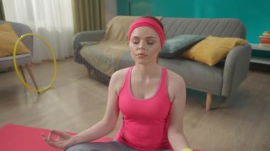 Genç ve çekici bir kadın lotus pozisyonunda oturur, gözleri kapalı, yavaş yavaş nefes alır ve verir. Evde yoga yapan kızıl saçlı kadının portresi. Spor konsepti. Yoga dersi. Yavaş