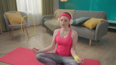 Kızıl saçlı, kulaklıklı genç bir kadın lotus pozisyonunda oturur, meditasyon yapar, yoga yapar. Spor giyimli bir kadının portresi oturma odasında yerde oturuyor. Rahatlama, meditasyon, yoga. Yavaş